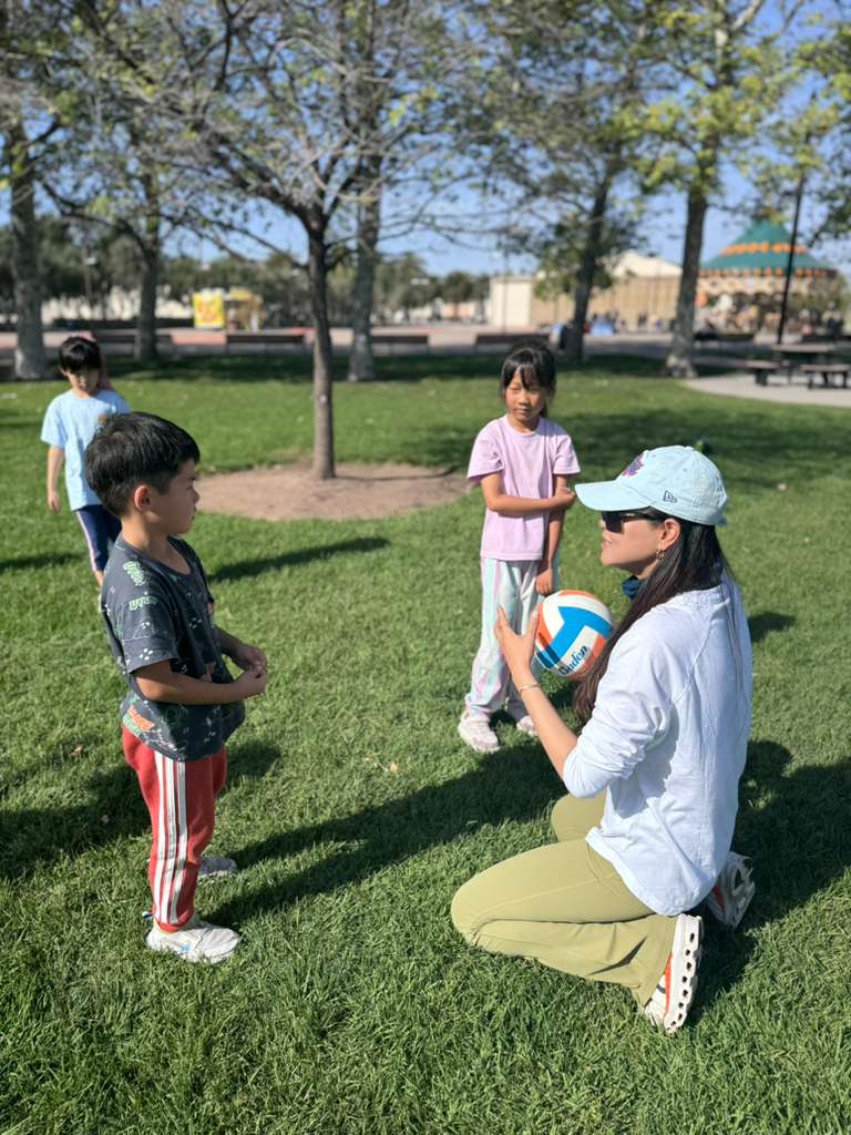 A coach kneels on the grass to meet a young boy at eye level, holding a volleyball, at a free class in Irvine.